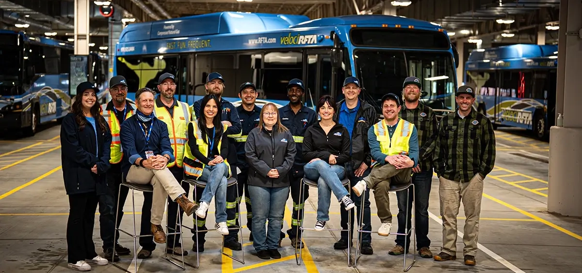 A group of diverse RFTA employees posed together in a RFTA bus maintenance facility in various RFTA branded wear.