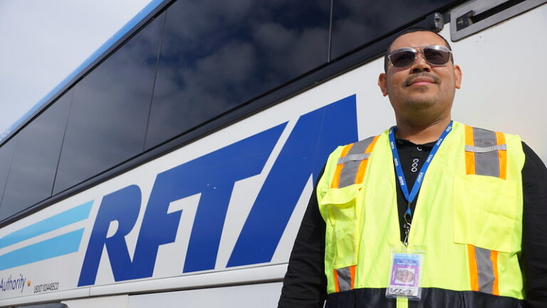 A RFTA bus operator stands next to a bus with a high visibility vest on